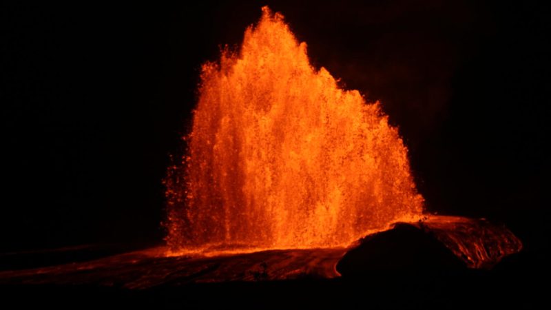 Night photo of a tall orange lava fountain at Kilauea's summit north vent, with glowing lava flows spreading across the dark ground in front of it.