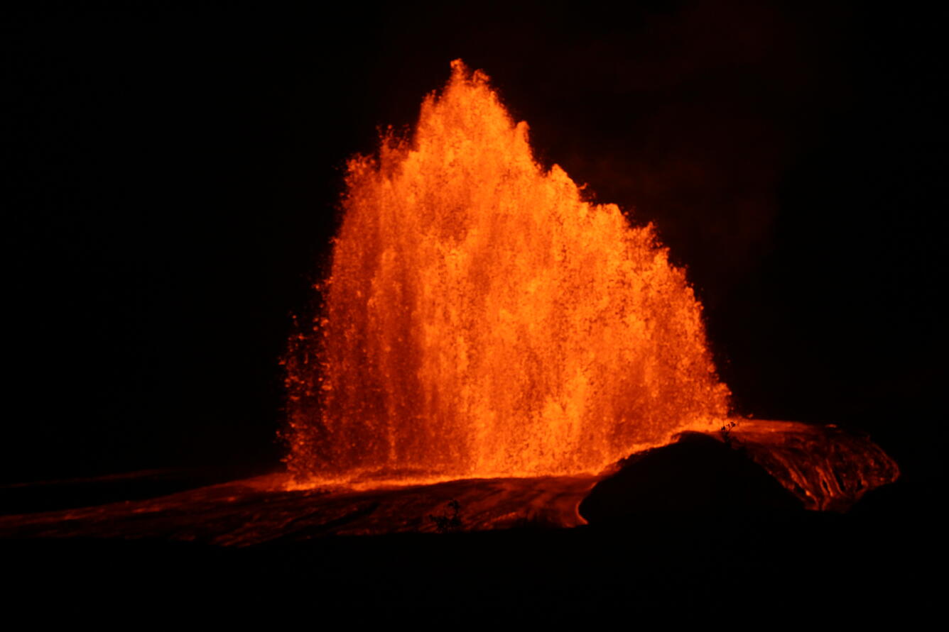 Night photo of a tall orange lava fountain at Kilauea's summit north vent, with glowing lava flows spreading across the dark ground in front of it.